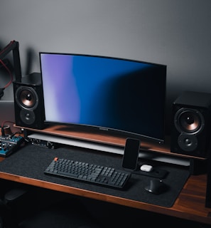 A modern gaming and streaming setup with a curved monitor placed centrally on a wooden desk. Surrounding the monitor are powerful speakers and a high-end PC tower with glowing fans. Various peripherals, including a mechanical keyboard, ergonomic mouse, and audio mixer, are neatly arranged. Overhead, a professional microphone and stand are ready for use, possibly for gaming commentary or podcasting.